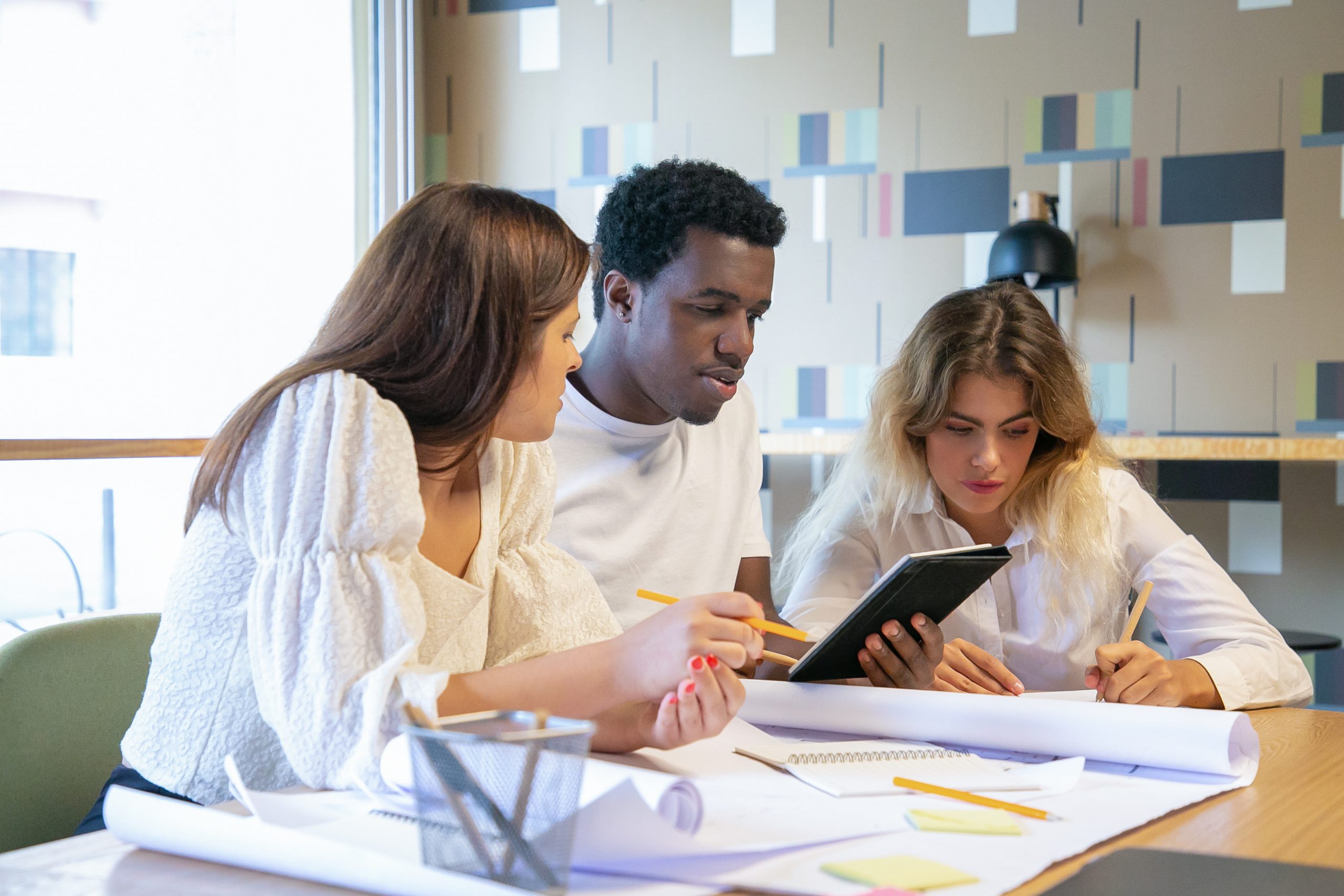 Creative professionals working on design project together. Professionals drawing in blueprints, writing notes in notebook, watching content on tablet. Medium shot. Creative job and teamwork concept