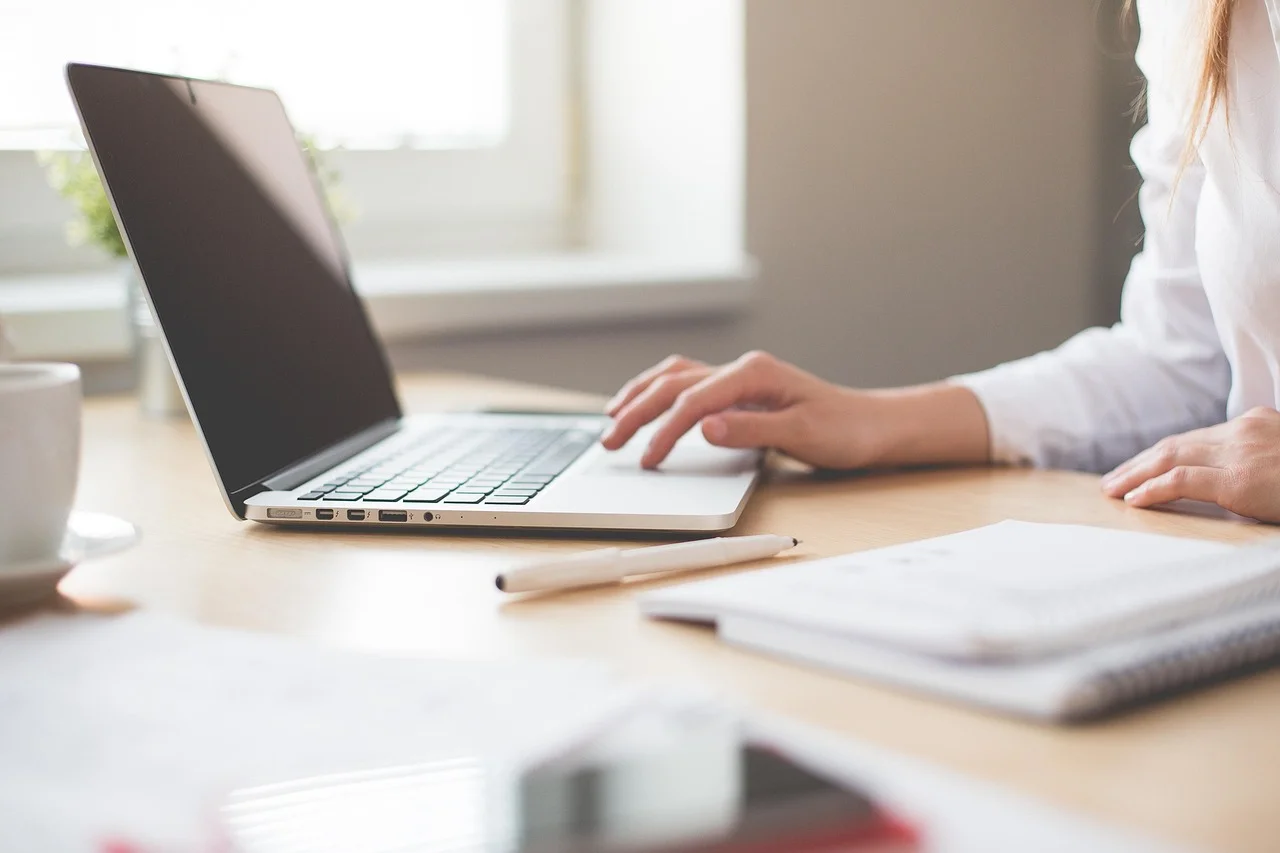 Person working on a laptop at a wooden desk next to a notebook and pen.